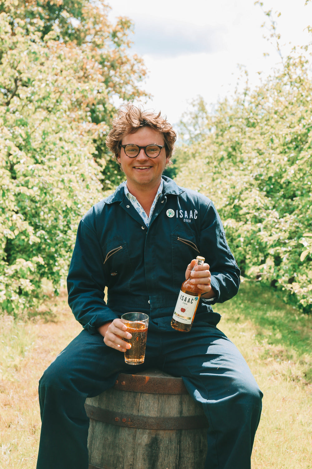 Rollo Wood of Isaac Cider seated among flowering orchard trees with a bottle and glass of cider.