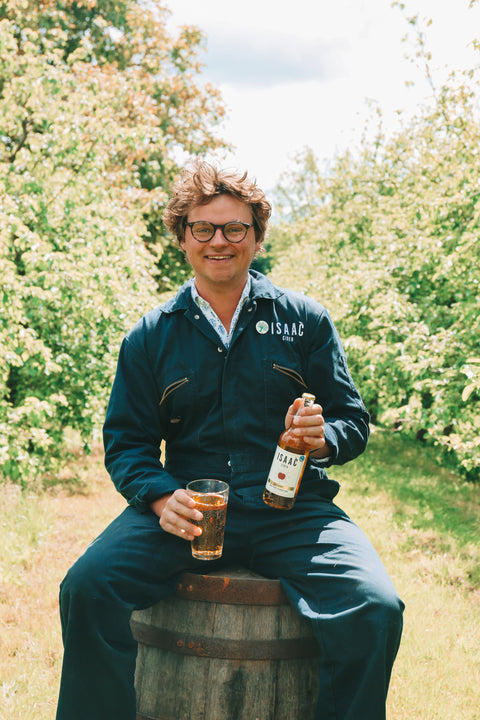 Rollo Wood of Isaac Cider seated among flowering orchard trees with a bottle and glass of cider.